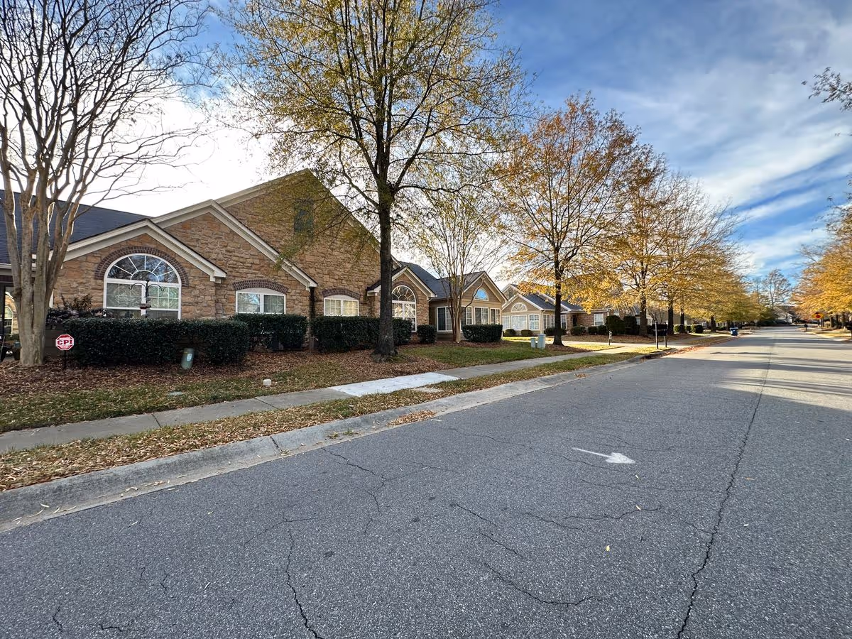 Street view of single-story brick senior living buildings with trees, a sidewalk, and a paved road under a partly cloudy sky.