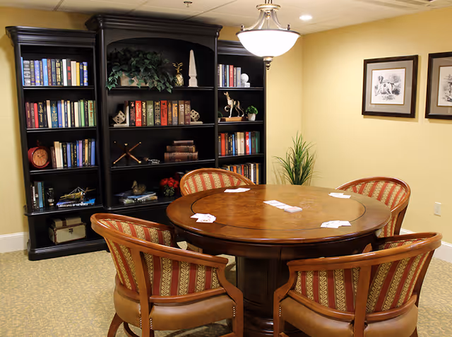 A cozy room with a round wooden table surrounded by five upholstered chairs with red and gold patterned fabric. On the table, there are playing cards spread out. Behind the table is a large black bookshelf filled with books and decorative items. The walls are painted light yellow, and there are two framed pictures hanging on the right wall. A ceiling light fixture hangs above the table, and a green plant is placed in the corner.