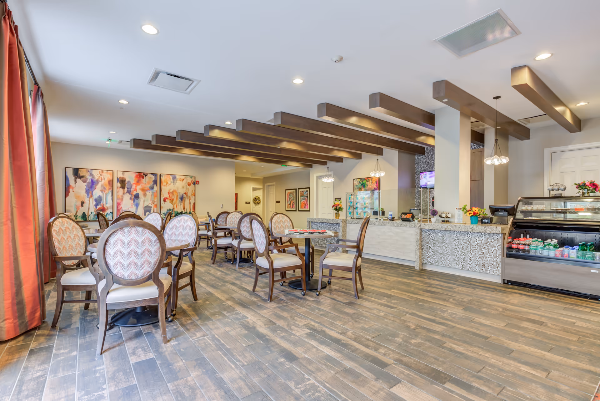 A bright and spacious dining area with multiple round tables and cushioned chairs arranged neatly. The room features wooden flooring, decorative ceiling beams, and large windows with red curtains allowing natural light to enter. On one side, there is a counter with a mosaic tile front and a refrigerated display case stocked with beverages. Colorful abstract paintings adorn the walls, and pendant lights hang from the ceiling.