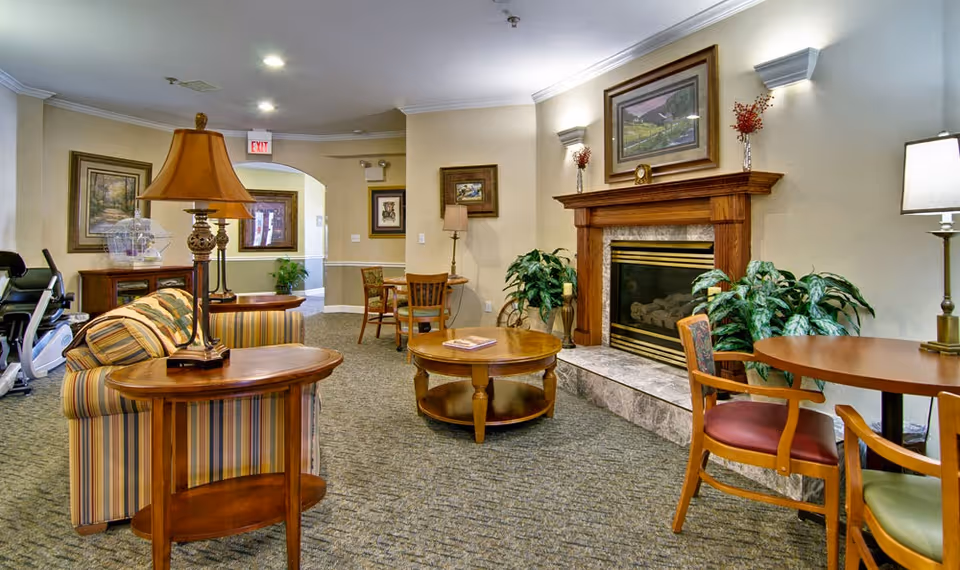 A cozy living room area in a senior living facility featuring a striped sofa, wooden side and coffee tables, a fireplace with a wooden mantle, framed artwork on the walls, potted plants, and several chairs around a round table. The room is warmly lit with lamps and wall sconces.