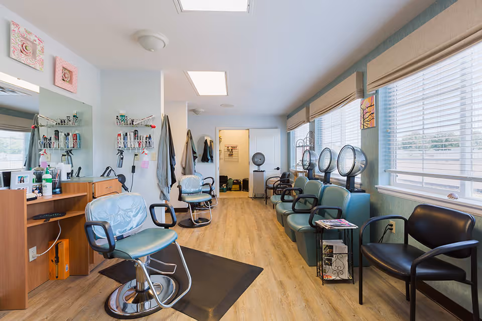 Interior view of a hair salon area in a senior living facility with salon chairs, hair dryers, mirrors, and shelves with hair products. The room has large windows with blinds and wooden flooring.