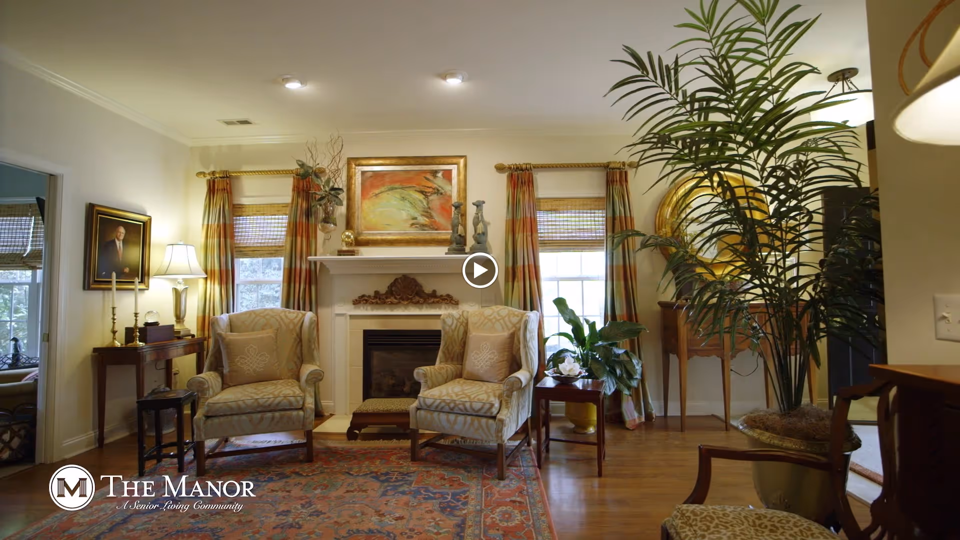 A cozy living room with two upholstered armchairs facing a fireplace. The fireplace mantel is decorated with art and sculptures. There are two windows with patterned curtains on either side of the fireplace. A large potted plant is on the right side of the room, and a small side table with a plant and flowers is between the chairs. The room has warm lighting and traditional decor, including a framed portrait on the left wall and a colorful area rug on the wooden floor.