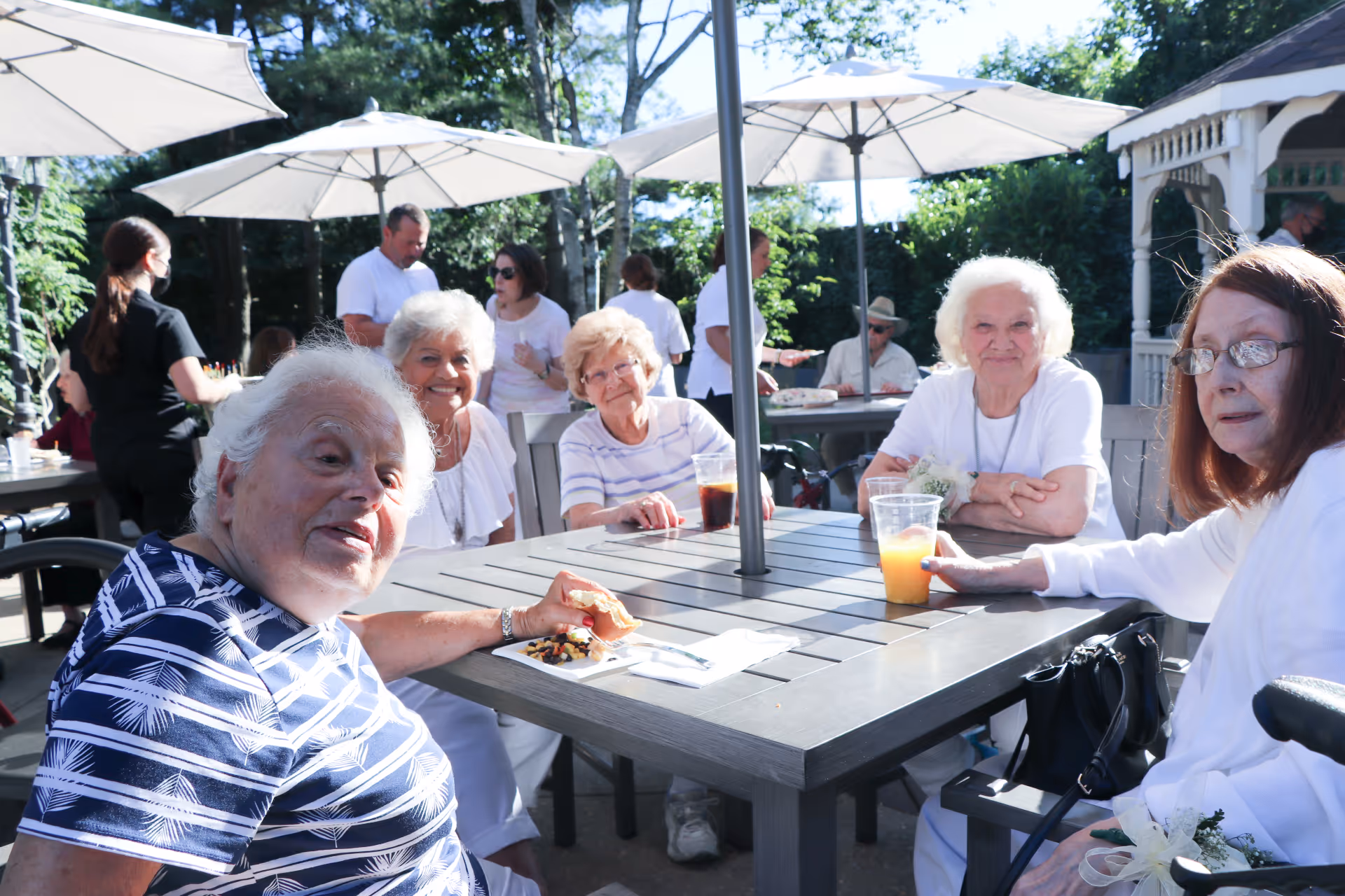 A group of elderly women sitting around a table outdoors under large white umbrellas, enjoying drinks and snacks on a sunny day. Other people are visible in the background, some standing and some seated, with greenery and a gazebo nearby.