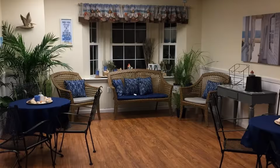 A cozy sitting area with wicker chairs and a loveseat adorned with blue cushions and pillows. There are two small round tables covered with dark blue tablecloths, each surrounded by black metal chairs. The room has wooden flooring, several potted plants, a window with a nautical-themed valance, and wall decorations including a framed quote and a bird sculpture.