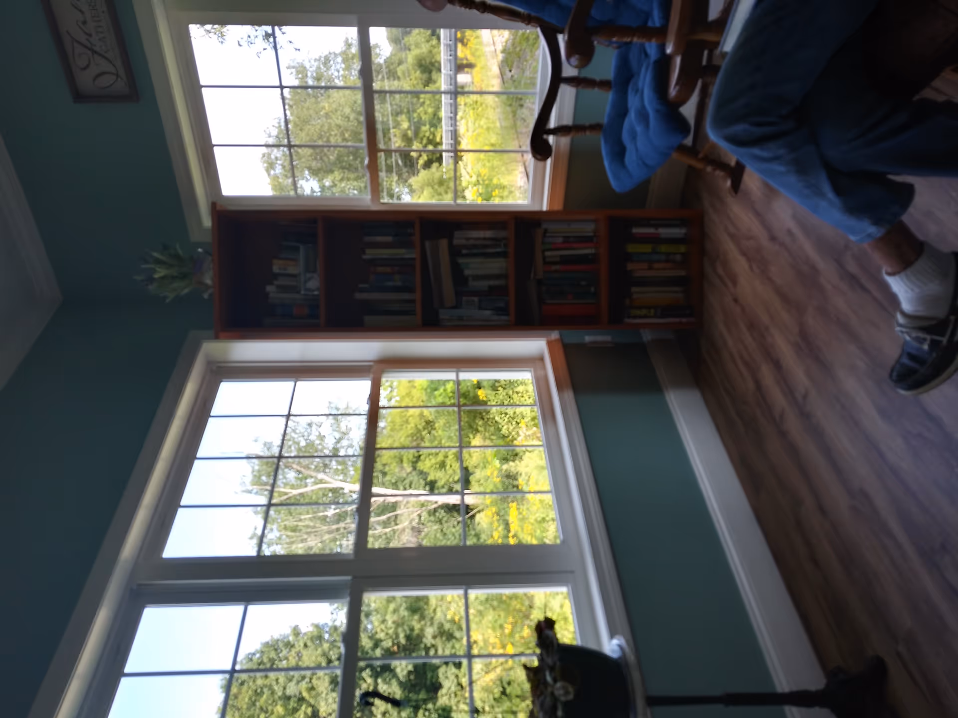 A cozy corner of a room with large windows showing a green outdoor view. There is a wooden bookshelf filled with books between the windows, a small round table with a potted plant on the left, and a wooden rocking chair with blue cushions on the right. A person wearing blue jeans, white socks, and black shoes is sitting in the chair with legs crossed. The walls are painted teal and the floor is wood.