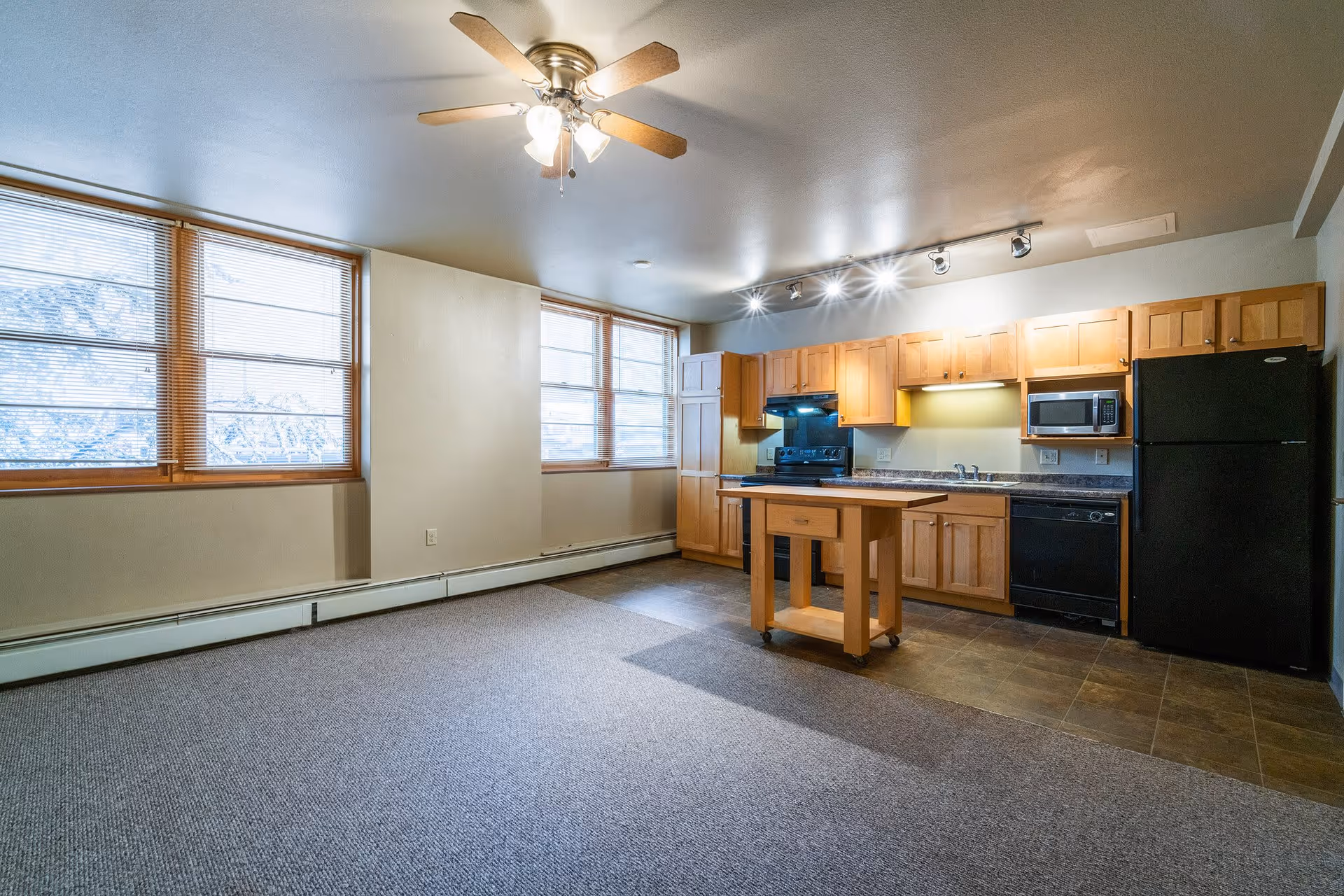 An empty senior living facility room with a kitchen area featuring wooden cabinets, a black refrigerator, microwave, stove, and dishwasher. The room has large windows with blinds, a ceiling fan with lights, and a small wooden kitchen island on wheels. The floor is a combination of carpet and tile.