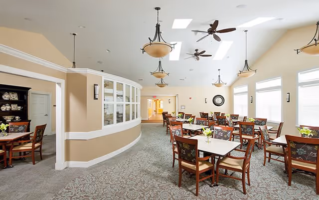 A bright dining room with multiple tables and chairs arranged neatly. The room has beige walls, large windows letting in natural light, ceiling fans, and hanging light fixtures. There is a carpeted floor with a patterned design and a curved wall with windows on the left side.