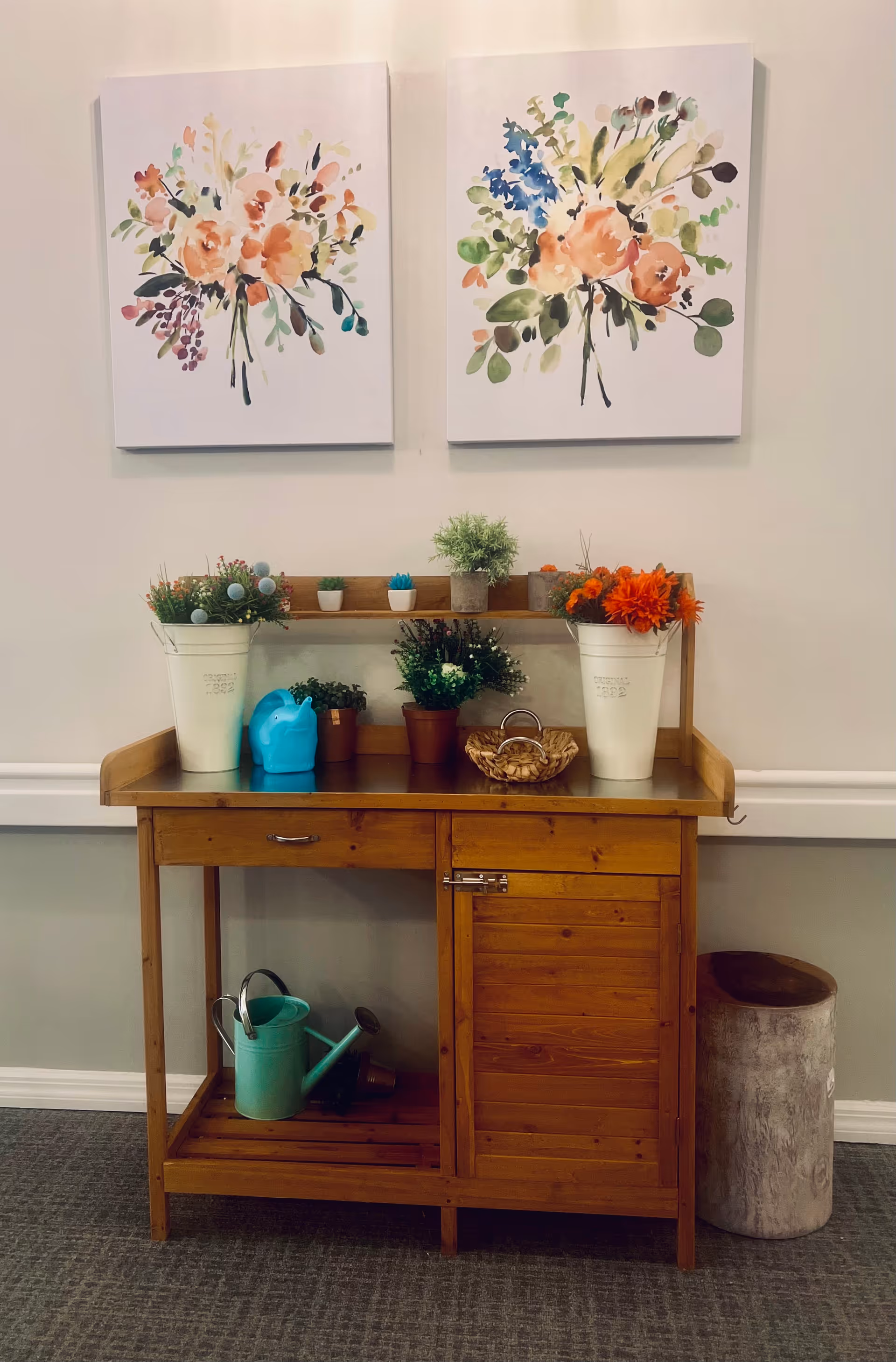 Wooden console table topped with potted plants, a blue watering can, and decorative items beneath two floral canvas paintings on a pale wall.
