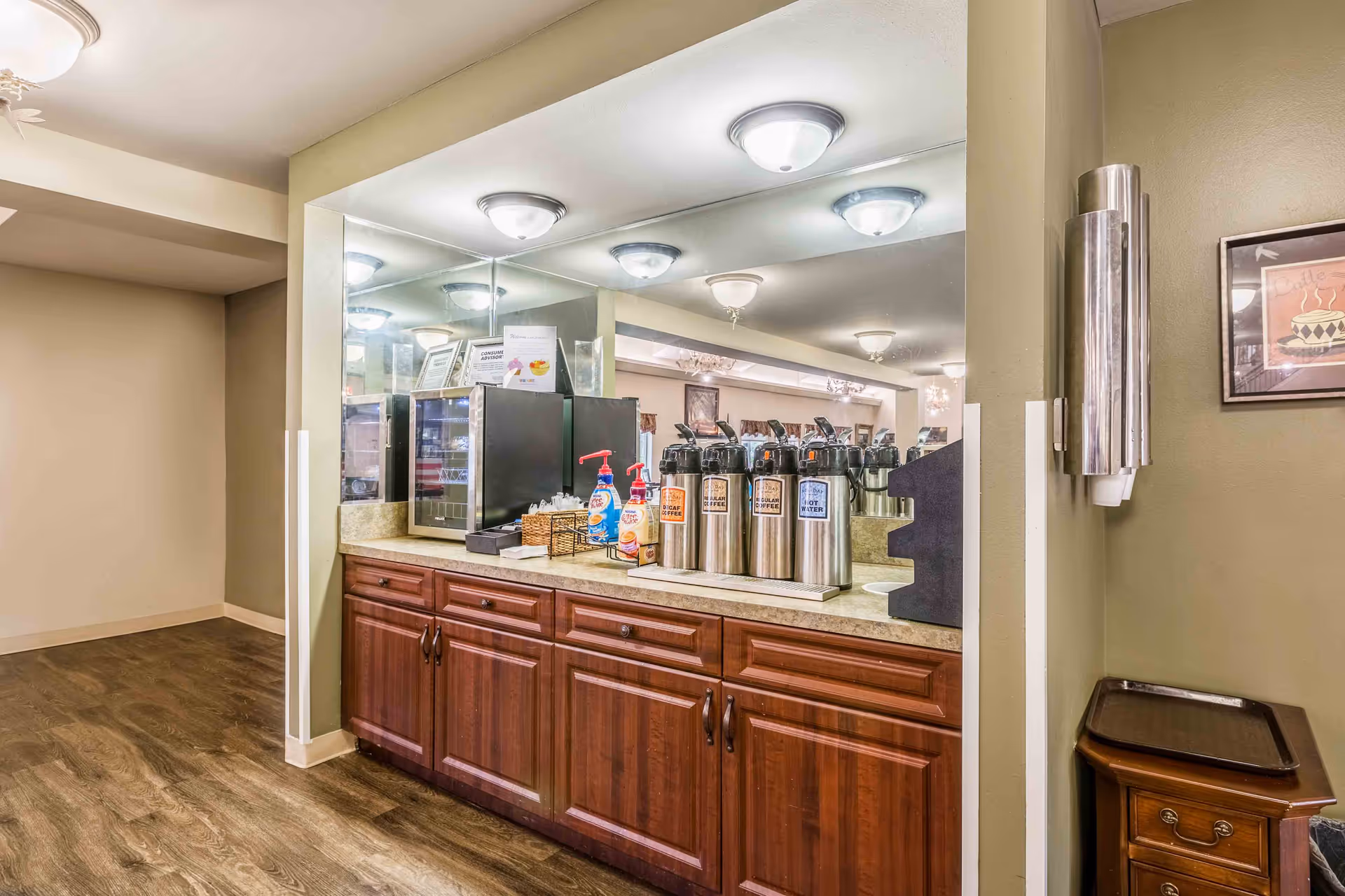 A beverage station with multiple stainless steel coffee dispensers labeled Decaf Coffee, Regular Coffee, and Hot Water on a countertop with wooden cabinets below. There are also bottles of coffee creamer and hand sanitizer, a small basket with packets, and a small refrigerator. The background features a large mirror reflecting ceiling lights and part of a dining area.
