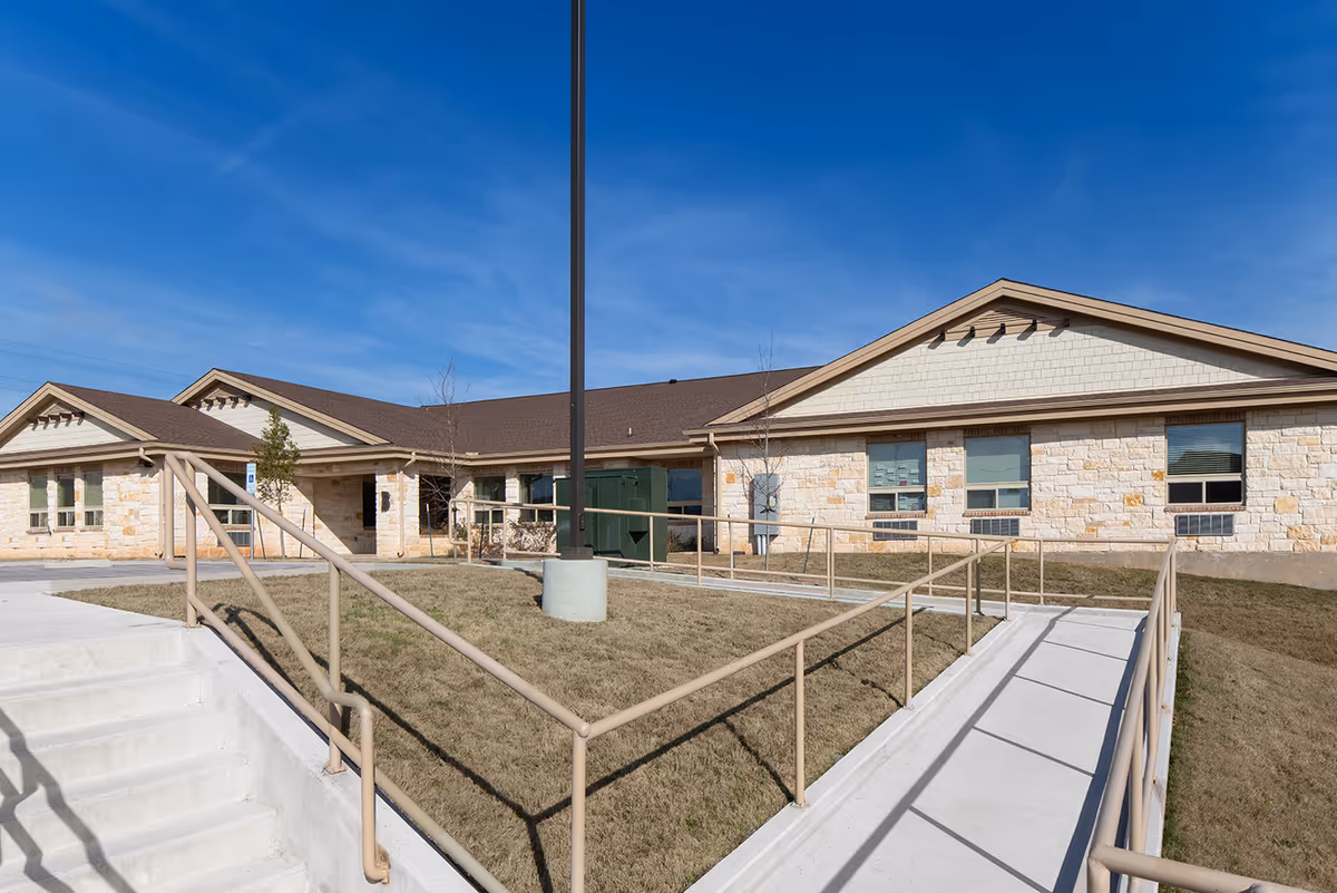 Exterior view of a single-story assisted living facility building with stone walls, multiple windows, a brown roof, a concrete ramp with handrails, and stairs leading to the entrance under a clear blue sky.