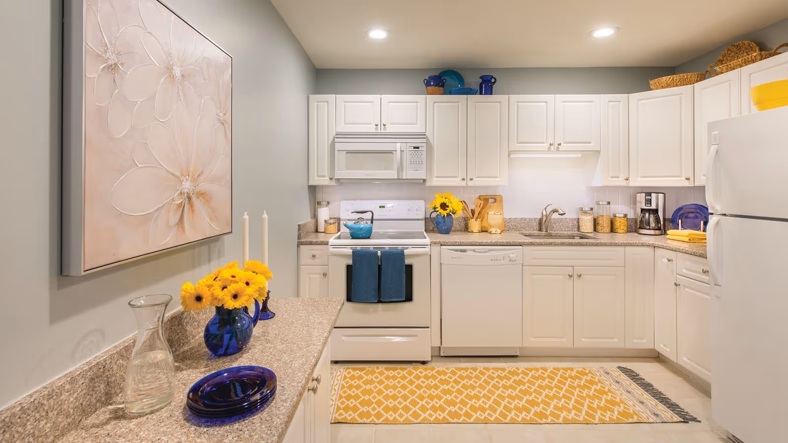 A bright and clean kitchen with white cabinets, a white stove, microwave, dishwasher, and refrigerator. The countertops are beige with a speckled pattern. There is a yellow patterned rug on the floor, a blue vase with yellow flowers, blue dishware, and various kitchen items neatly arranged. A large floral artwork hangs on the left wall.