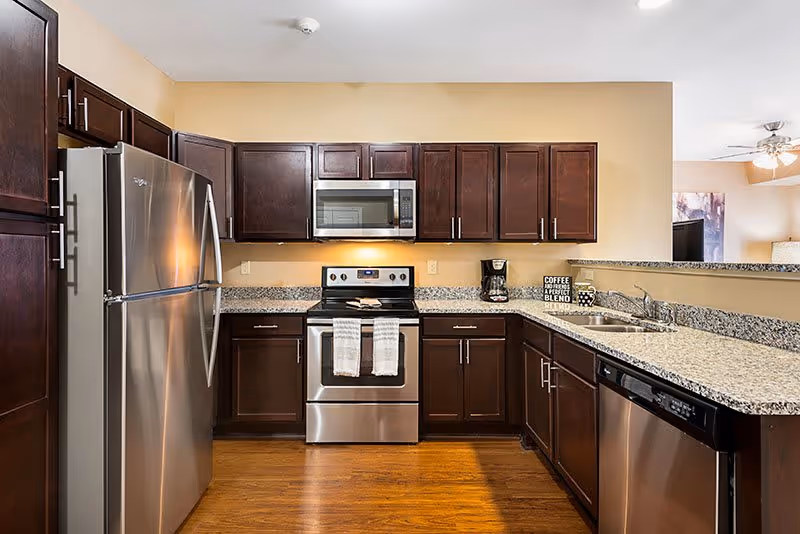 Modern kitchen with dark wood cabinets, stainless steel refrigerator, stove, microwave, dishwasher, and granite countertops. The kitchen has a coffee maker and a decorative sign on the counter. The floor is wooden and the kitchen opens to a living area with a ceiling fan and lamp visible in the background.
