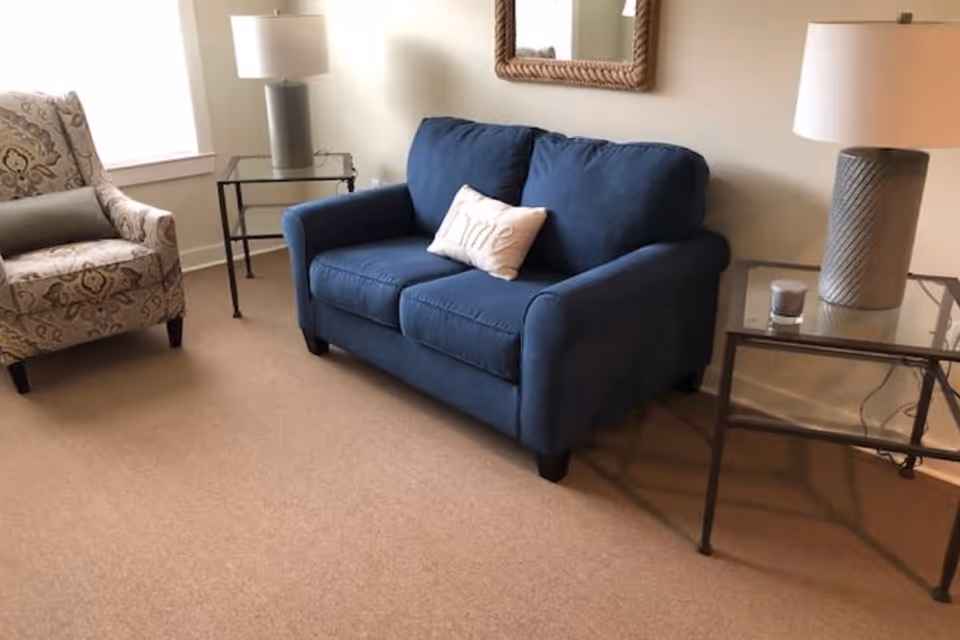 Small living room with a blue loveseat, patterned armchair, glass side tables, lamps, and a wall mirror.