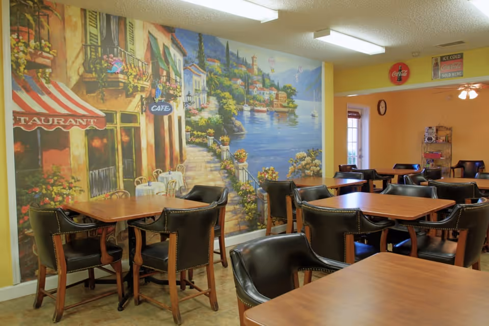 Dining area with several wooden tables and black leather chairs. The walls are painted yellow and feature a large mural depicting a scenic waterfront village with colorful buildings, flowers, and boats on the water. There are also vintage Coca-Cola signs on the wall in the background.