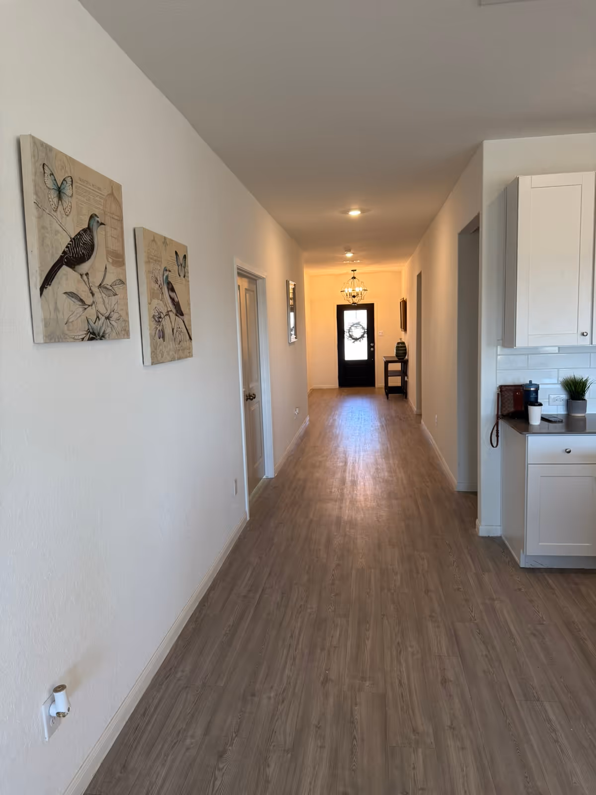 A long hallway with light wood flooring and white walls. On the left wall, there are two framed pictures of birds and butterflies. The hallway leads to a black door with a wreath, and a small table with a green vase is placed near the door. On the right side, part of a white kitchen cabinet and countertop with some items on it are visible.