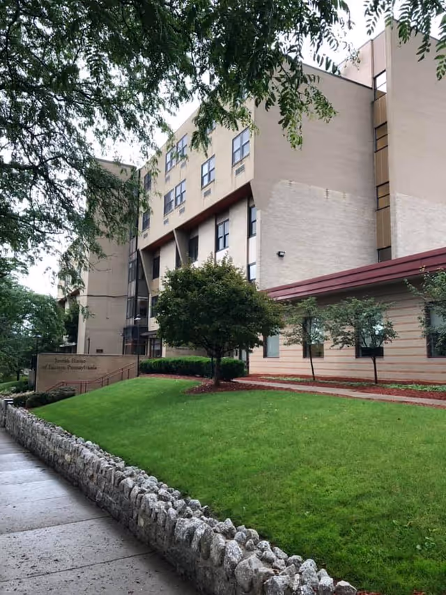 Exterior view of a multi-story beige building with several windows, surrounded by green grass, trees, and a stone wall along a sidewalk. The sky is overcast and the building appears to be a senior living or healthcare facility.