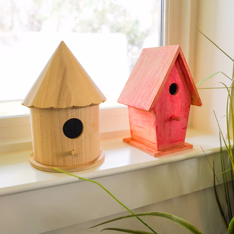 Two decorative wooden birdhouses placed on a white windowsill. One birdhouse is natural wood color with a conical roof, and the other is painted pink with a pitched roof. Green plant leaves are partially visible in the foreground.
