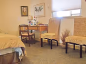 Small furnished bedroom with a bed, wooden desk and chair, two woven side chairs and a window with a shade.