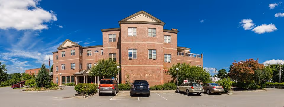 Front exterior view of a three-story brick building with multiple windows, a small covered entrance, and a parking lot with several cars parked. The sky is clear and blue with some clouds, and there are trees and shrubs around the building.