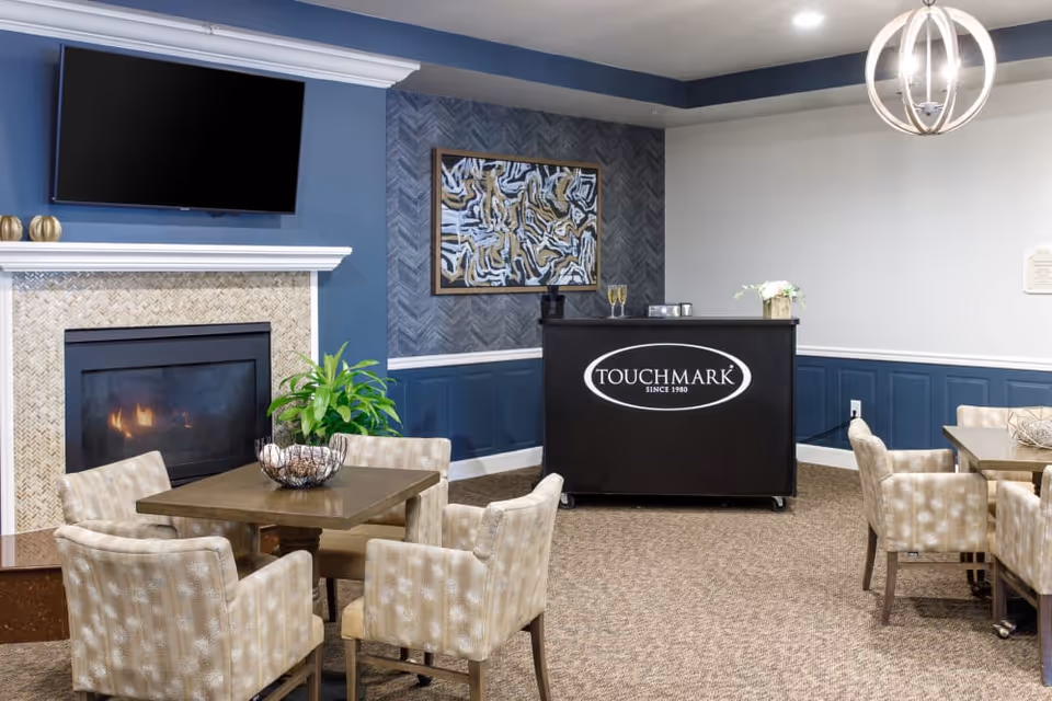 Community lounge/dining area with tables and upholstered chairs, a fireplace and wall-mounted TV, and a cart branded "Touchmark".