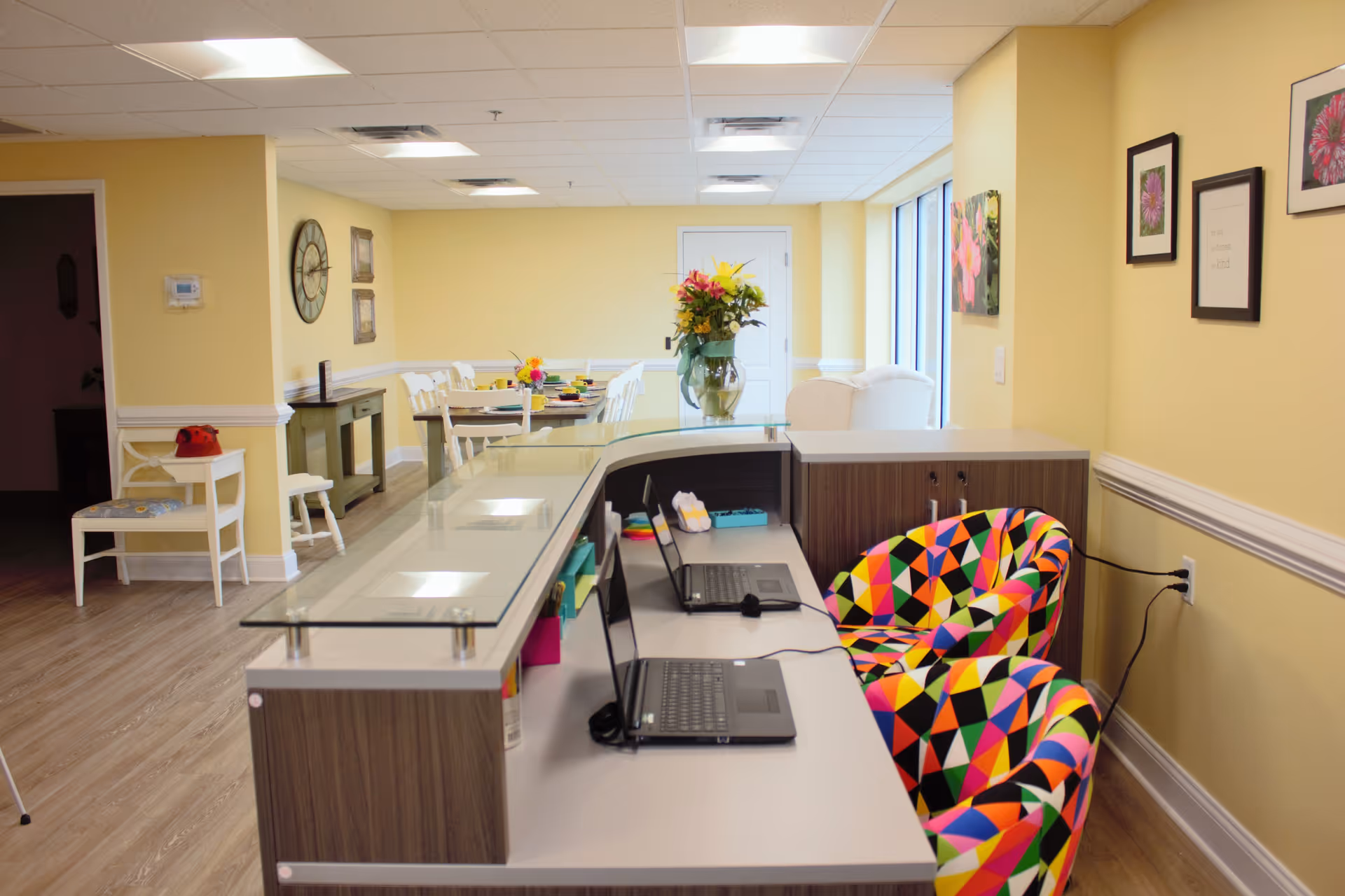 Interior view of a senior living facility reception or common area with a long desk featuring two colorful geometric-patterned chairs and two laptops. In the background, there is a dining table set with colorful dishes and a vase of flowers. The walls are painted yellow and decorated with framed pictures and a large clock.