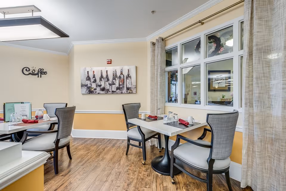 A cozy dining area with two tables, each set with glasses, cups, and red napkins. The room has wooden flooring, light yellow walls with white trim, and a large window with beige curtains. There is a wall decoration with wine bottles and glasses, and a decorative sign that says 'Coffee'.