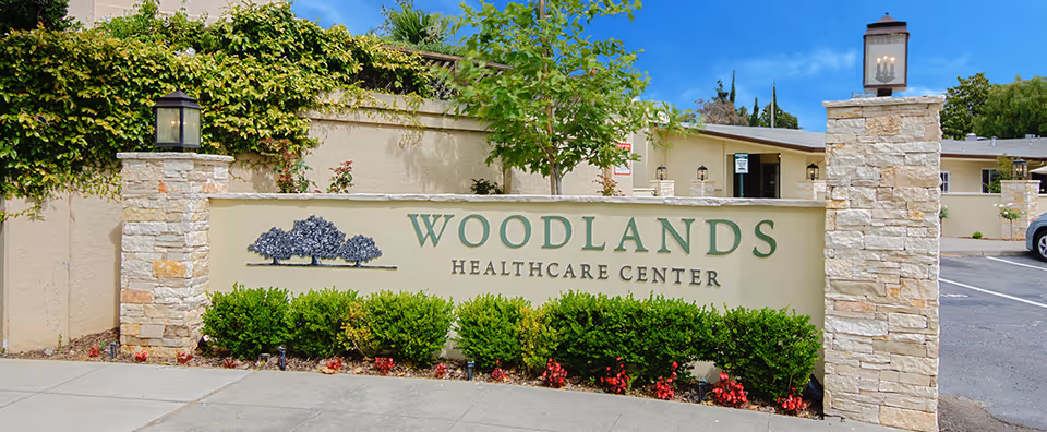 Entrance sign reading "Woodlands Healthcare Center" mounted on a low stone wall with shrubs, lantern pillars, and the facility building behind.