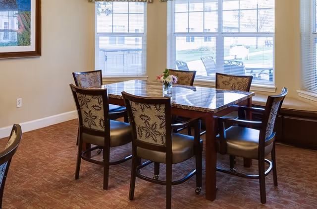 A dining area with a square table featuring a marble top surrounded by six wooden chairs with patterned upholstery. The room has large windows with blinds, allowing natural light to brighten the space. A small vase with flowers is placed on the table. The walls are light-colored, and there is a framed picture hanging on one wall.