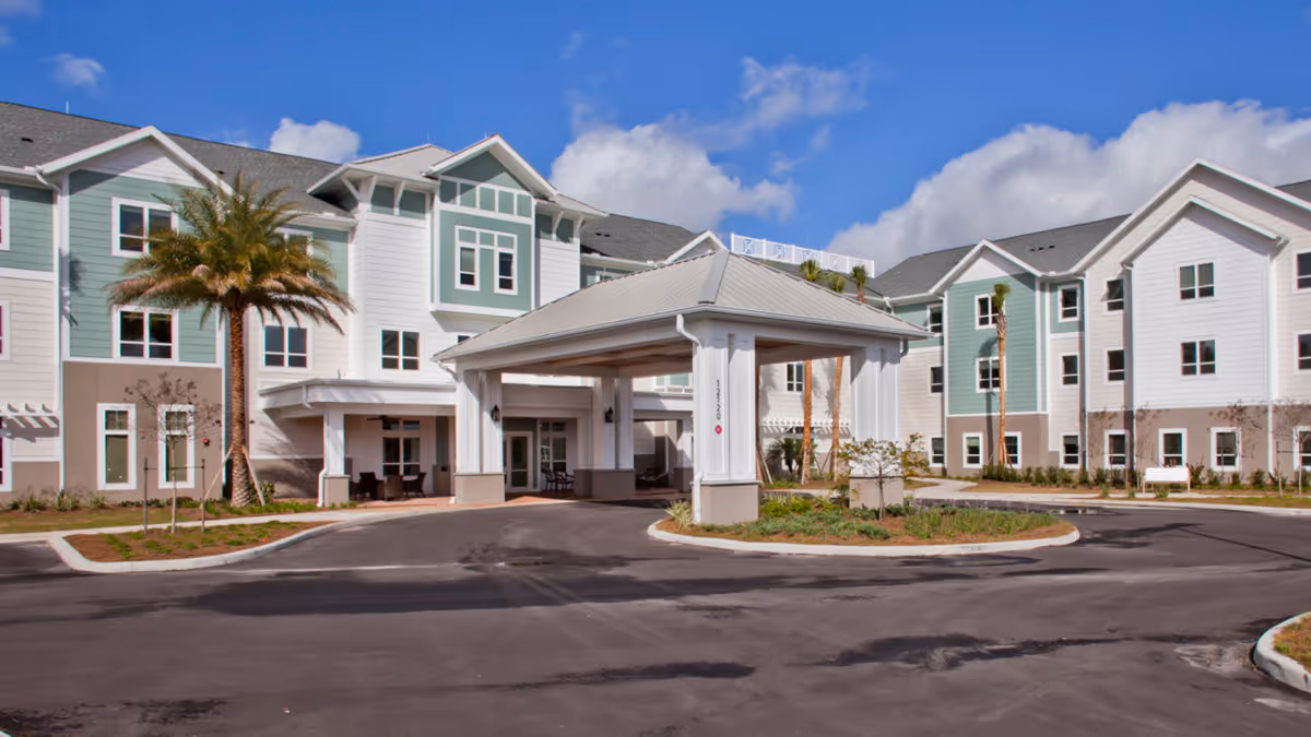 Front entrance and porte-cochere of a three-story senior living building with pastel siding, palm trees, and a circular driveway.