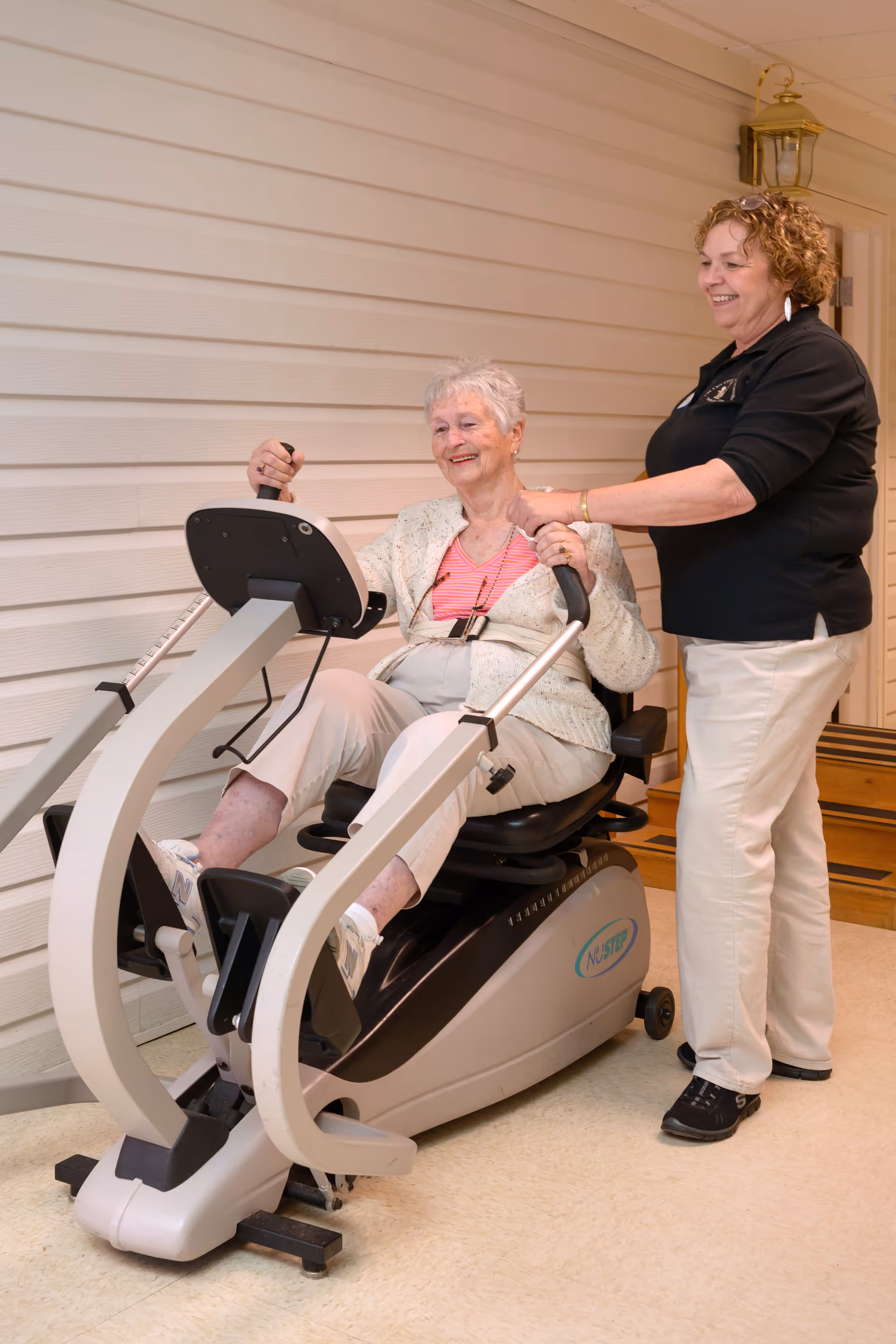 An elderly woman is seated on a NuStep exercise machine, smiling while holding the handles. A caregiver stands beside her, providing support and encouragement. They are indoors against a beige paneled wall.