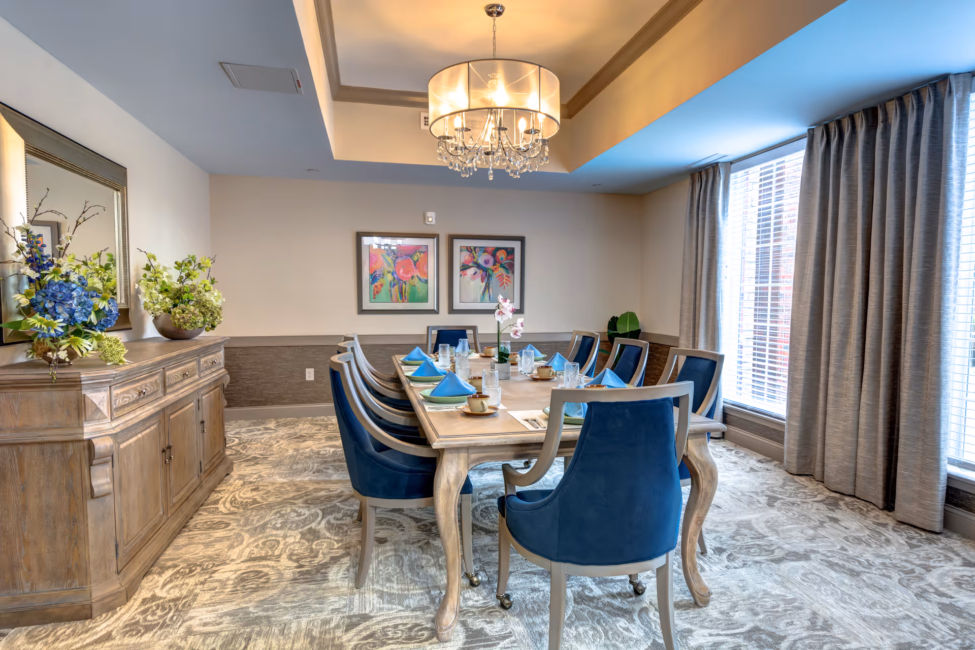 A dining room with a long wooden table set for a meal, featuring blue upholstered chairs, blue folded napkins, glassware, and cups. The room has a patterned carpet, large windows with gray curtains, a chandelier hanging from the ceiling, a wooden sideboard with flower arrangements, and two colorful framed paintings on the wall.