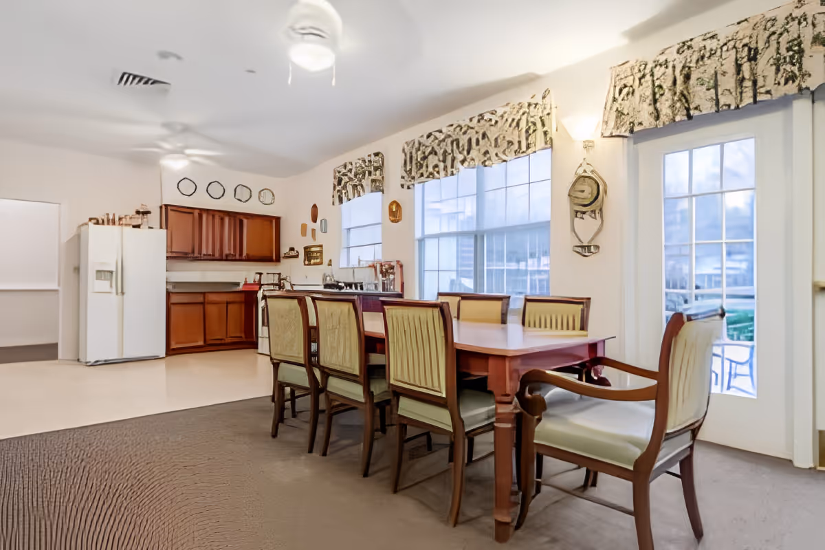 Dining area with a long wooden table and chairs next to a small kitchen and large windows.