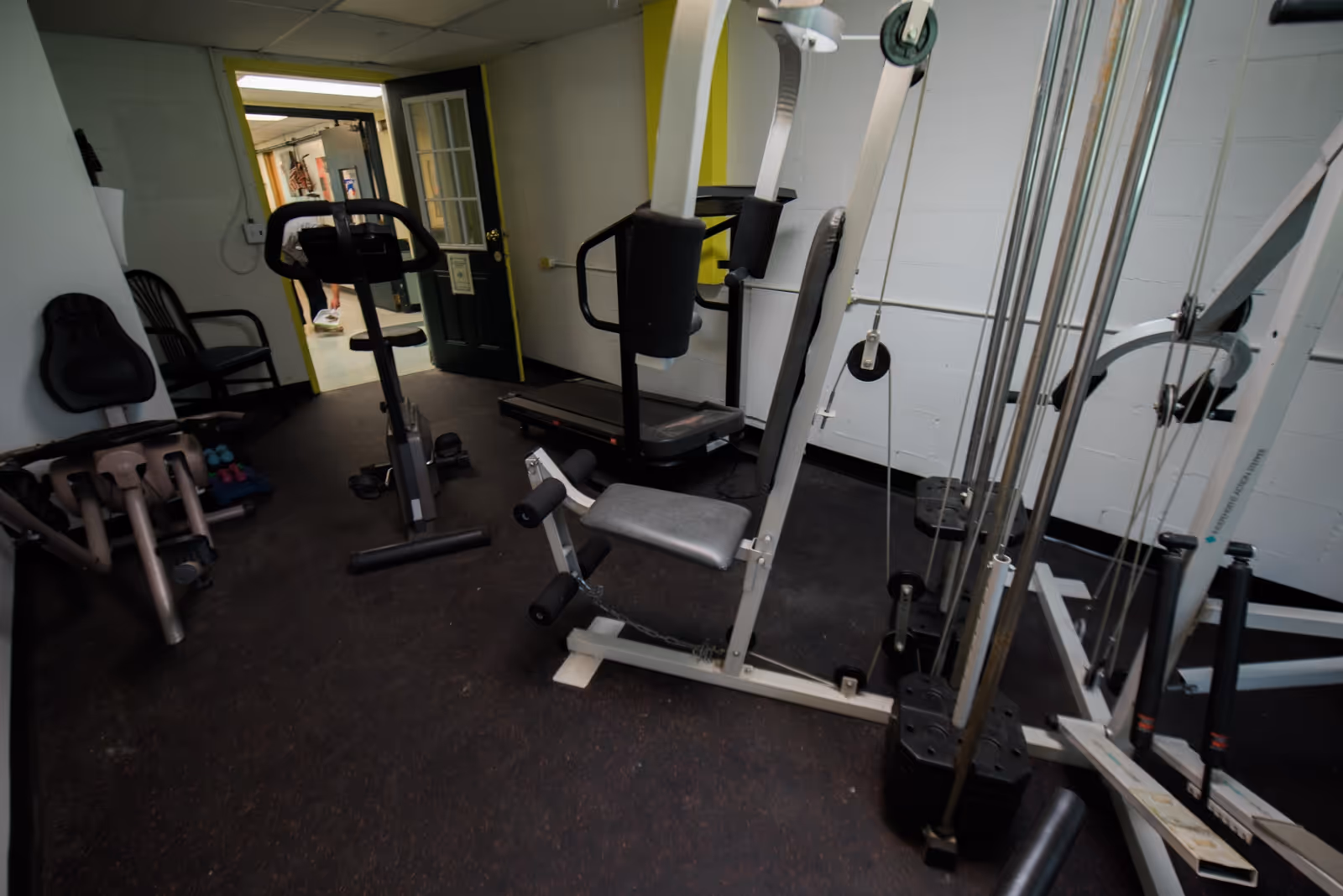 Small fitness room with various exercise equipment including a stationary bike, treadmill, weight machine, and dumbbells. The room has a dark floor and white walls, with an open door leading to a hallway.