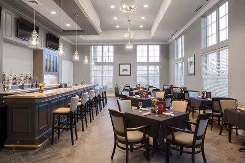 A spacious dining area with multiple tables covered in dark tablecloths, each set with plates, glasses, and napkins. There are cushioned chairs around the tables. To the left, there is a long bar with high stools and pendant lights hanging above. Large windows allow natural light to fill the room, and the ceiling has recessed lighting and decorative molding.