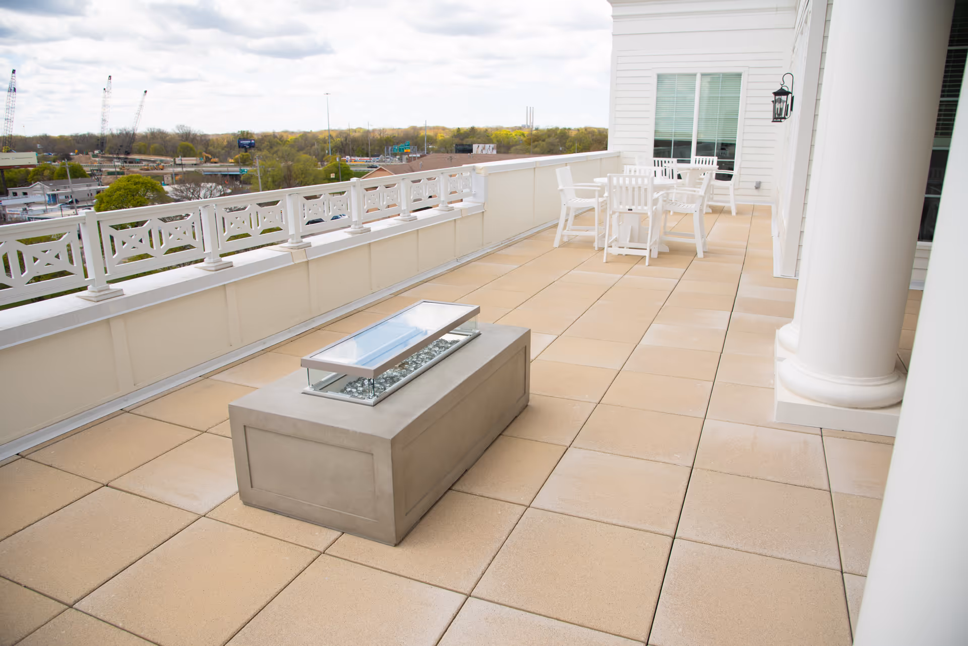 Outdoor patio area with beige tiled flooring, a rectangular fire pit with glass cover, white railing, white chairs and tables, and large white columns attached to a building. Trees and a partly cloudy sky are visible in the background.