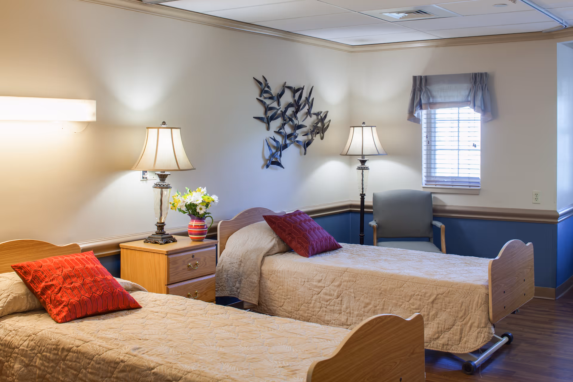 A cozy bedroom with two single beds, each with beige quilts and red pillows. Between the beds is a wooden nightstand with a decorative lamp and a vase of flowers. A metal wall art piece depicting birds is mounted on the wall above the nightstand. There is a window with blinds and a valance, and a cushioned armchair is placed near the window. The room has soft lighting and a warm, inviting atmosphere.