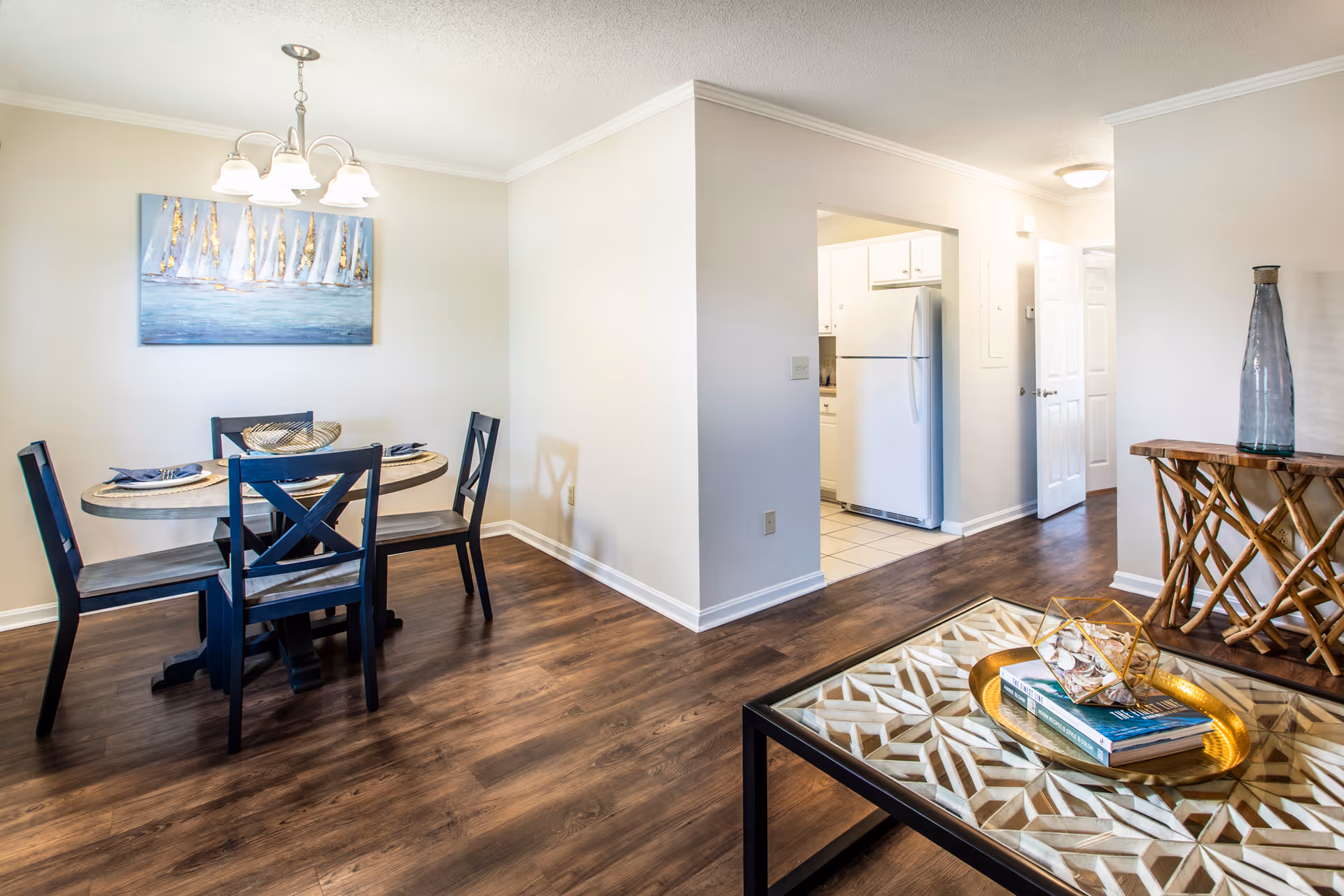Interior view of a senior living facility showing a dining area with a round table and four chairs, a decorative painting of sailboats on the wall, and a living room area with a glass coffee table holding books and decorative items. The kitchen with a white refrigerator is visible through an open doorway, and the floor is covered with wood-style flooring.