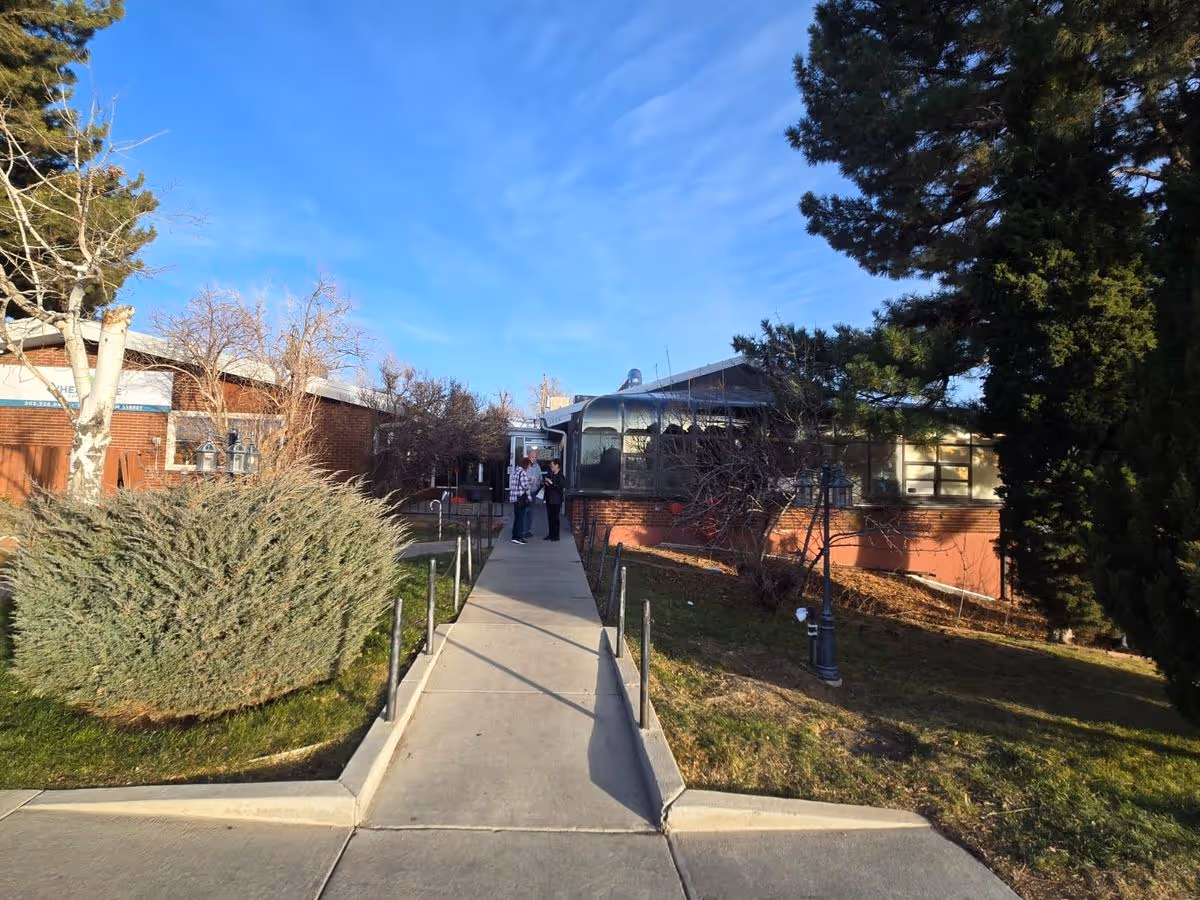 Concrete walkway leading to the entrance of a one-story brick care facility flanked by shrubs and trees under a blue sky.