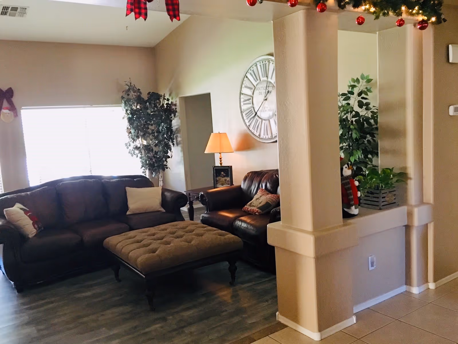 A cozy living room with a dark brown leather sofa and loveseat, a large tufted ottoman in the center, a side table with a lamp, a large wall clock, and decorative plants. The room is decorated with Christmas ornaments and garlands.