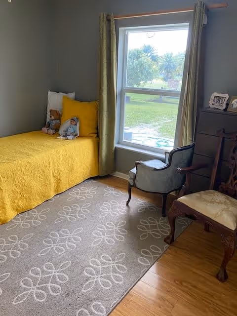 A cozy bedroom with a single bed covered in a yellow quilt, two dolls placed on the bed, a window with green curtains letting in natural light, a patterned beige rug on the wooden floor, and two chairs near a dark wooden dresser with framed photos on top.