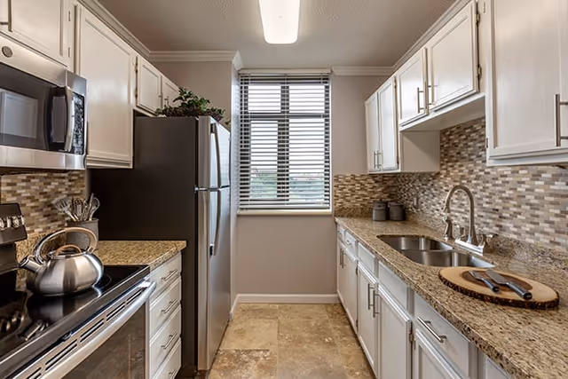 A modern kitchen with white cabinets, granite countertops, a stainless steel refrigerator, microwave, and stove. There is a window with blinds at the end of the kitchen, mosaic tile backsplash, and a double sink with a faucet. A cutting board with a knife is placed on the countertop.