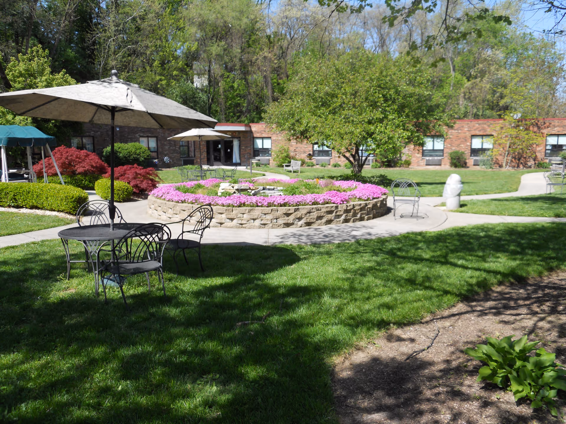 Outdoor garden area at Gardens of Oakley featuring a circular raised flower bed with pink flowers, surrounded by paved walkways, metal tables and chairs with umbrellas, green grass, trees, and a brick building in the background.