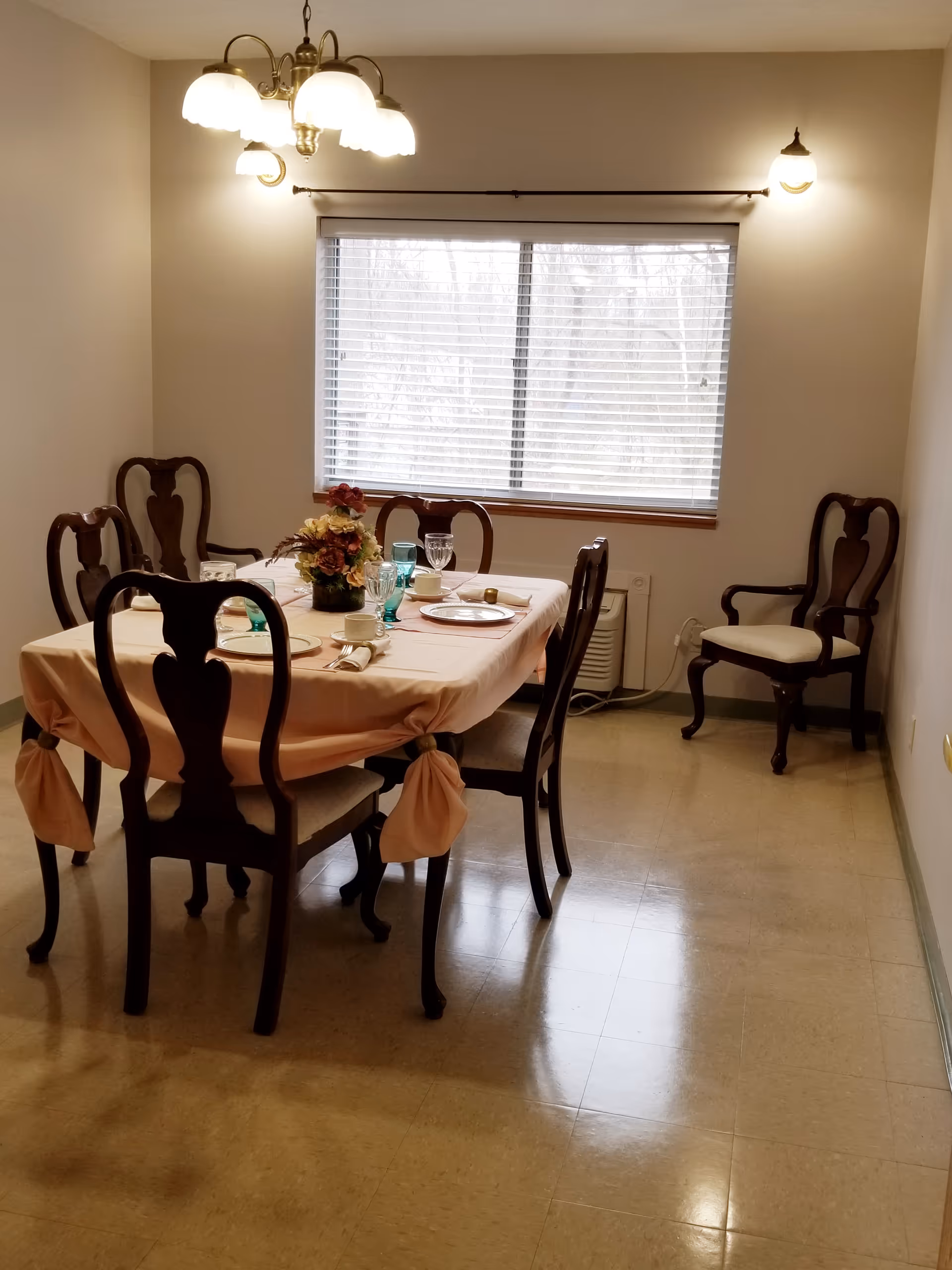 A dining room with a rectangular table covered with a peach-colored tablecloth tied at the corners. The table is set with plates, cups, glasses, and a floral centerpiece. There are six wooden chairs around the table and one additional chair against the wall. A window with blinds is on the far wall, and two wall-mounted lights and a ceiling light fixture illuminate the room.