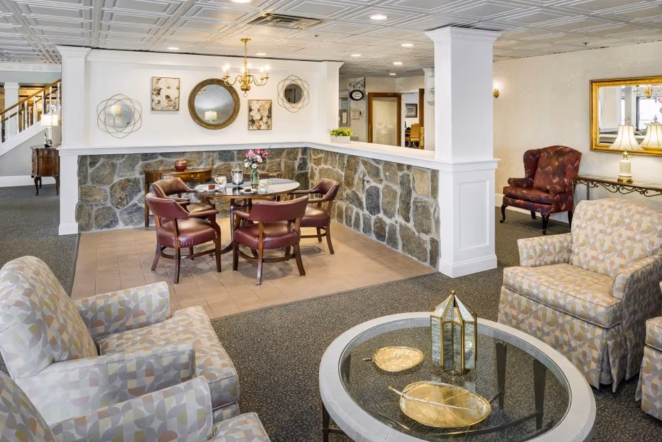 A bright senior living facility common room with upholstered armchairs around a glass coffee table and a small dining area with a round table and leather chairs.