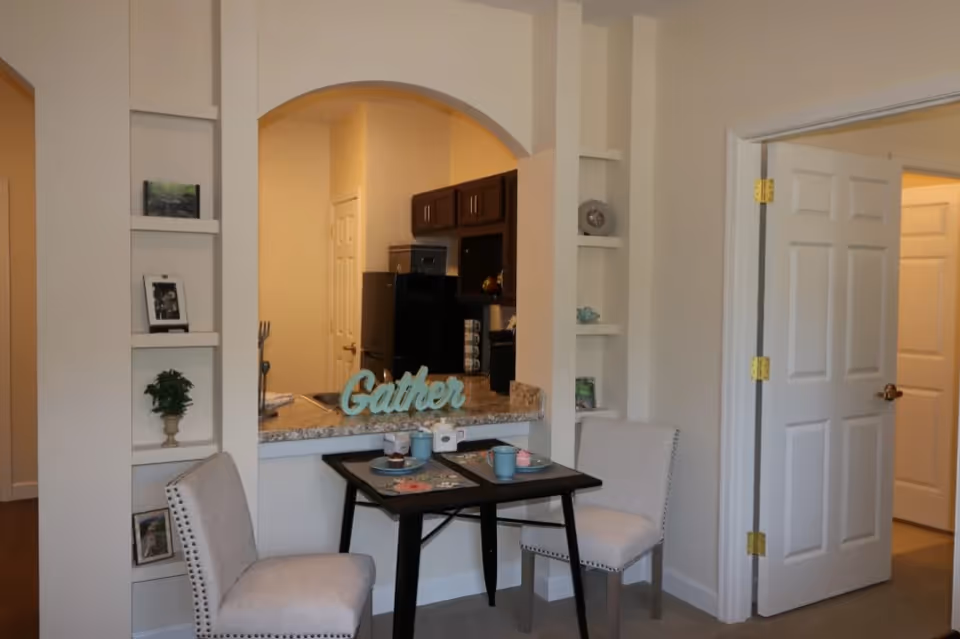 Cozy dining nook with two upholstered chairs and a small table set for tea in front of a kitchen pass-through with a 'Gather' sign on the counter.