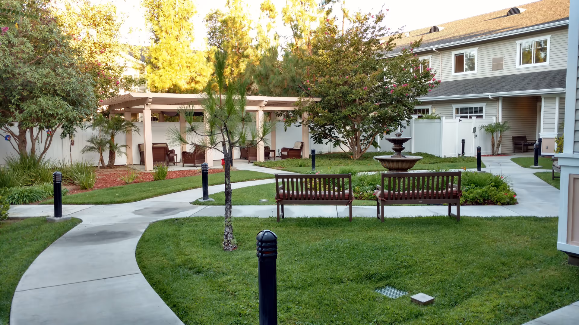 Outdoor courtyard area with green grass, a concrete pathway, two wooden benches facing a tiered water fountain, trees, and a covered seating area with chairs. Residential-style buildings surround the courtyard.