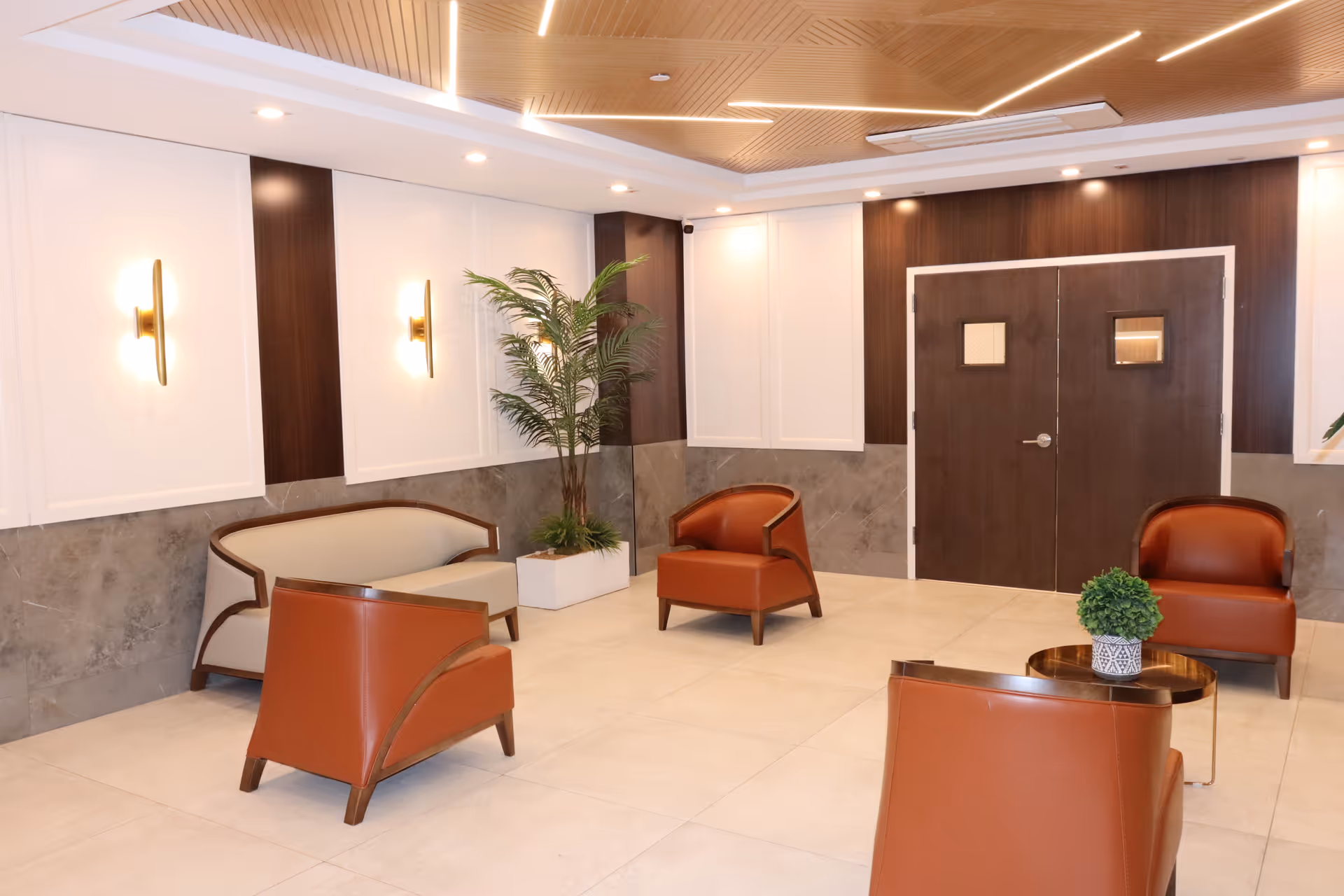 A modern, well-lit waiting or lounge area with four brown leather armchairs arranged around a small round table with a potted plant. There is a beige loveseat against the wall, a tall potted plant in the corner, and double wooden doors with small windows. The walls are decorated with white paneling and dark wood accents, and the ceiling has recessed lighting and a wooden design.