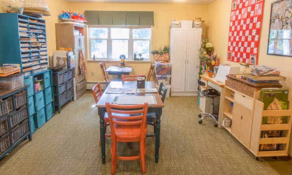 Bright interior activity room with tables and colorful chairs, storage cabinets and craft supplies beneath a window.