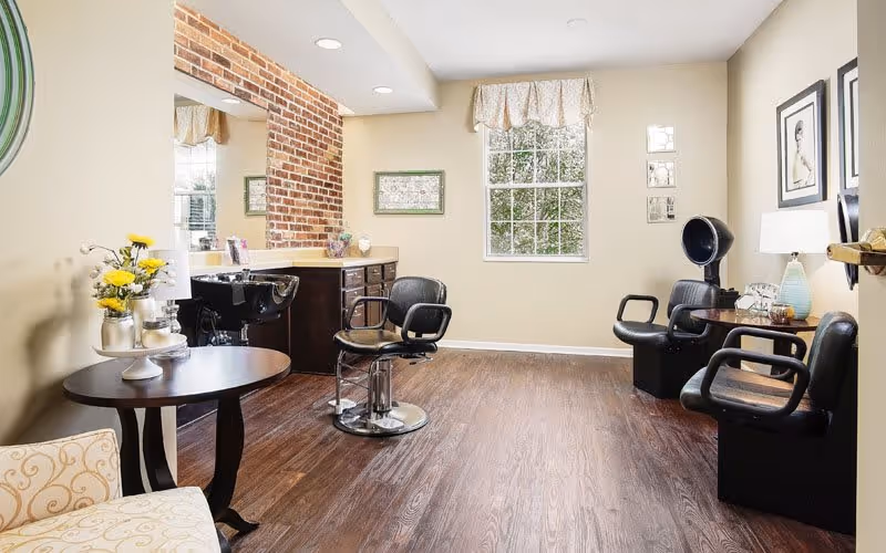 A bright and clean hair salon area with wooden flooring, beige walls, and a large window letting in natural light. The room features two black salon chairs, a hair washing station with a black sink, a small round table with yellow flowers in a vase, and framed pictures on the walls.