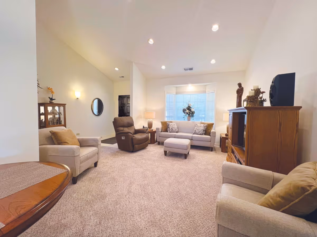 A cozy living room with beige carpet and neutral-colored furniture including a sofa, two armchairs, and a brown recliner. The room features a bay window with blinds and a floral arrangement on the windowsill. There is a wooden entertainment center with decorative items and a speaker, a round wooden table partially visible in the foreground, and soft lighting from wall sconces and table lamps.